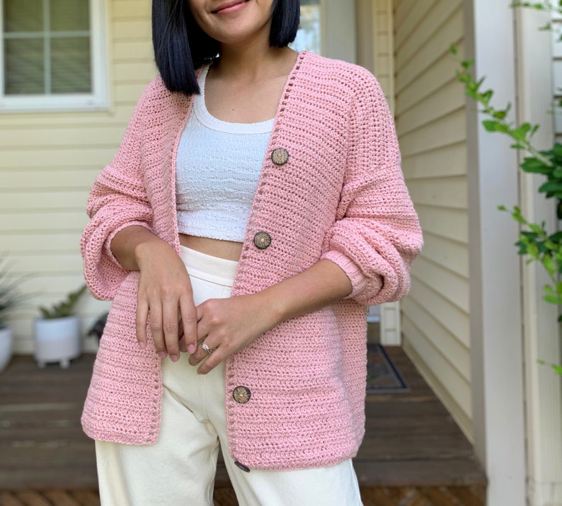 May include: A pink crocheted cardigan with wooden buttons. The cardigan has a v-neck and long sleeves.