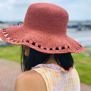 May include: A terracotta-colored crochet sun hat with a wide brim and decorative cut-out edging. The hat has a rounded crown and is made of a textured yarn. The hat is designed to provide shade from the sun.
