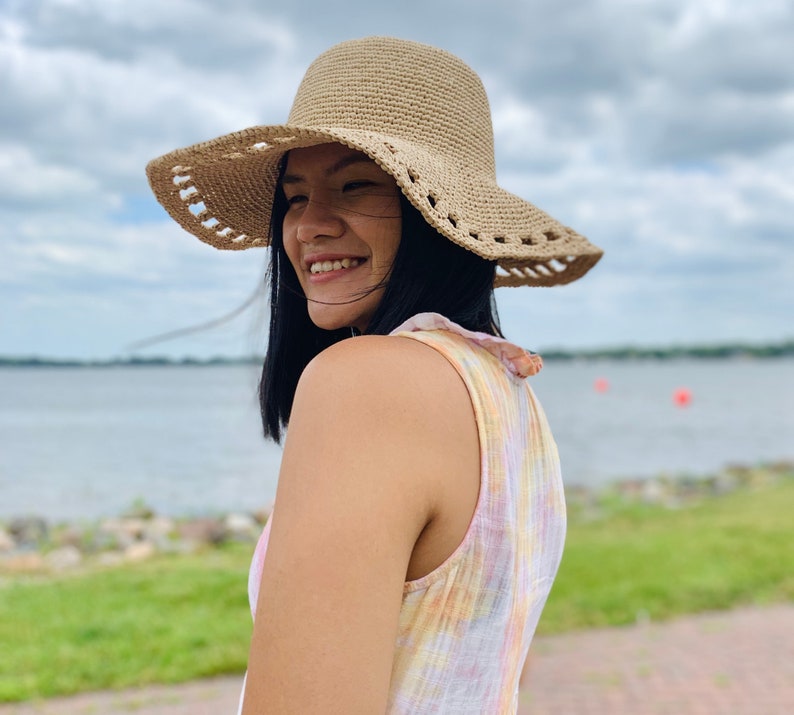 May include: A beige sun hat with a wide brim and decorative cut-out detail. The hat is made of woven material and is worn outdoors on a cloudy day. The hat's brim measures approximately 15 inches in diameter.