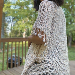 May include: A white crochet cardigan with a speckled pattern and a fringe trim. The cardigan has a loose fit and a V-neck.