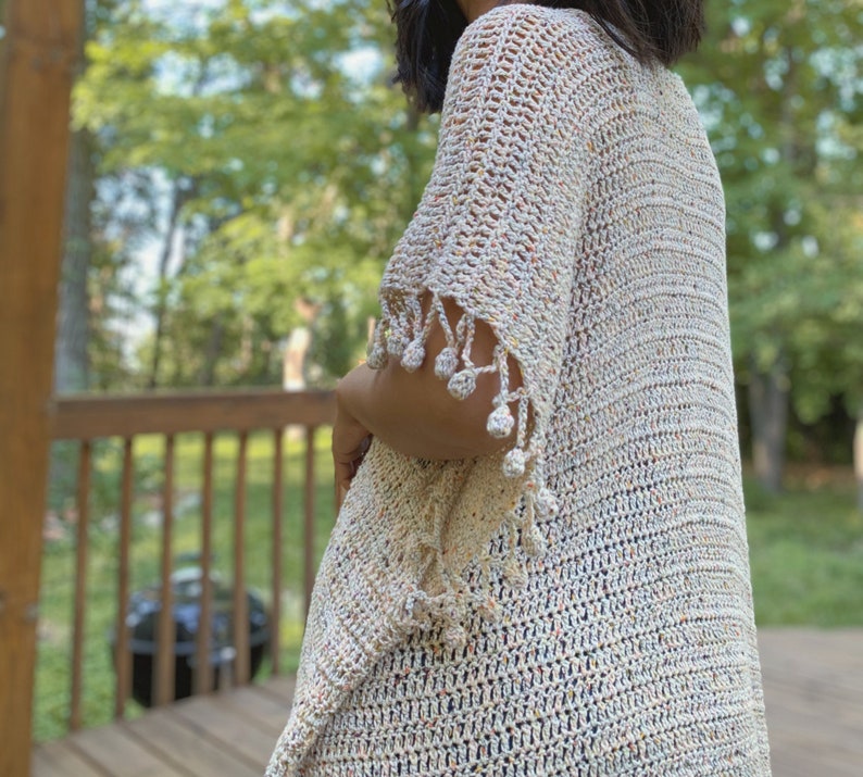 May include: A white crochet cardigan with a speckled pattern and a fringe trim. The cardigan has a loose fit and a V-neck.