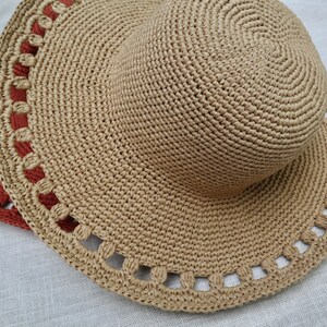 May include: A close-up of two woven straw hats. The primary hat is beige with a wide brim and a decorative scalloped edge. A second hat, partially visible, is a darker reddish-brown color.