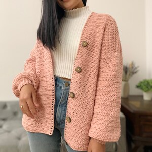 May include: A pink crocheted cardigan with a V-neck and button closure. The cardigan has long sleeves and a relaxed fit.