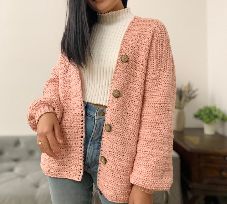 May include: A pink crocheted cardigan with a V-neck and button closure. The cardigan has long sleeves and a relaxed fit.