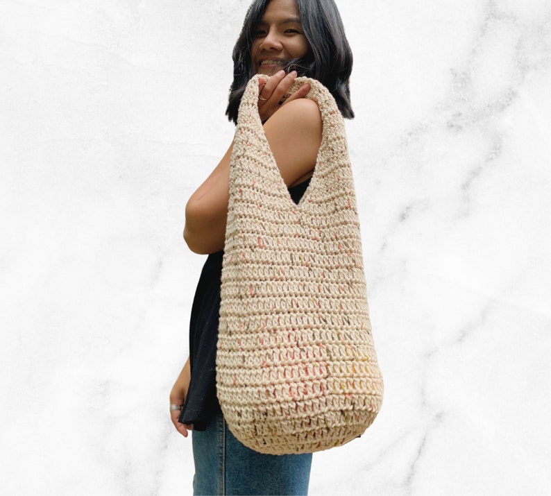 May include: A beige crocheted tote bag with a speckled pattern. The bag has a wide, open top and a long strap.