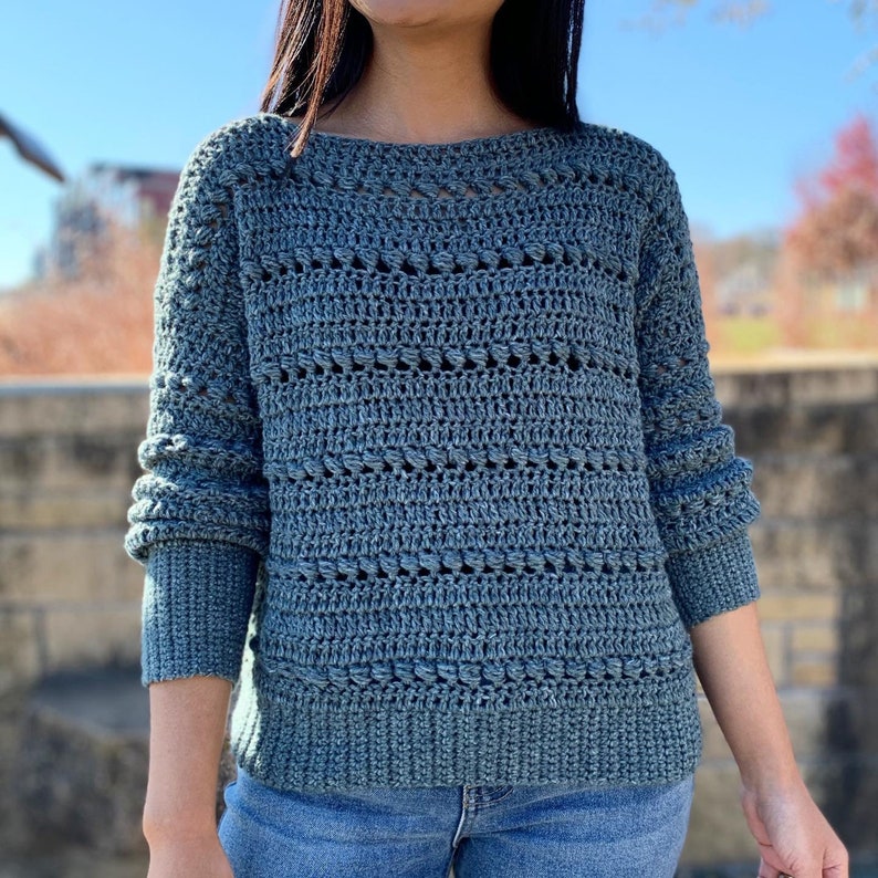 May include: A blue crocheted sweater with a loose fit and a textured design. The sweater has a boat neck and long sleeves.