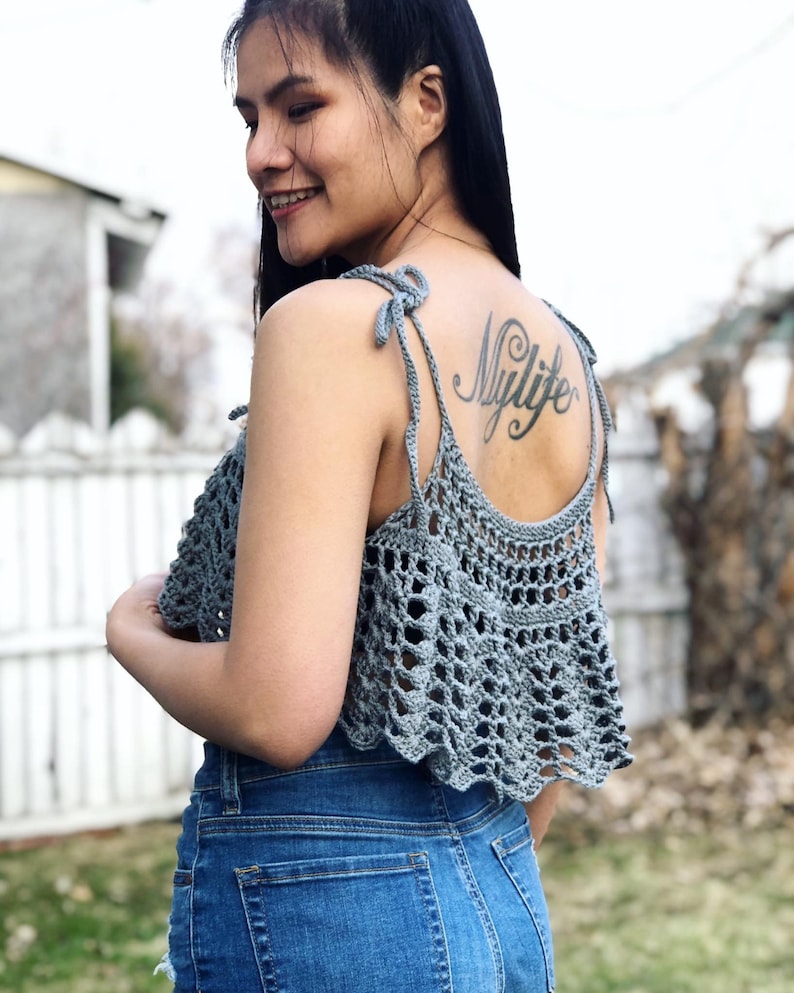 May include: A grey crochet top with a tie-up halter neckline. The top has a scalloped hem and a delicate openwork pattern. The model is wearing the top with blue denim shorts and has a tattoo on her back that says "Mylife".