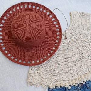 May include: A rust-colored crochet sun hat with a wide brim and decorative cutouts. A cream-colored crochet top with a round neckline and a denim skirt are also visible. A necklace with a pendant is also present.