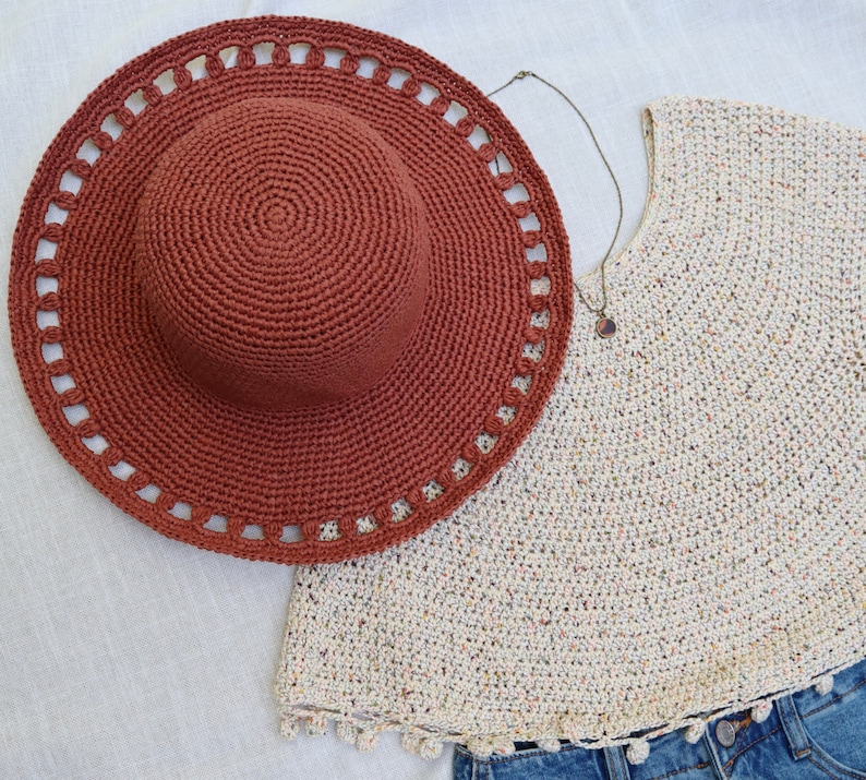 May include: A rust-colored crochet sun hat with a wide brim and decorative cutouts. A cream-colored crochet top with a round neckline and a denim skirt are also visible. A necklace with a pendant is also present.