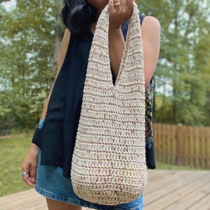 May include: A crocheted tote bag with a cream base and multi-colored flecks. The bag has a wide, v-shaped opening and a long strap.