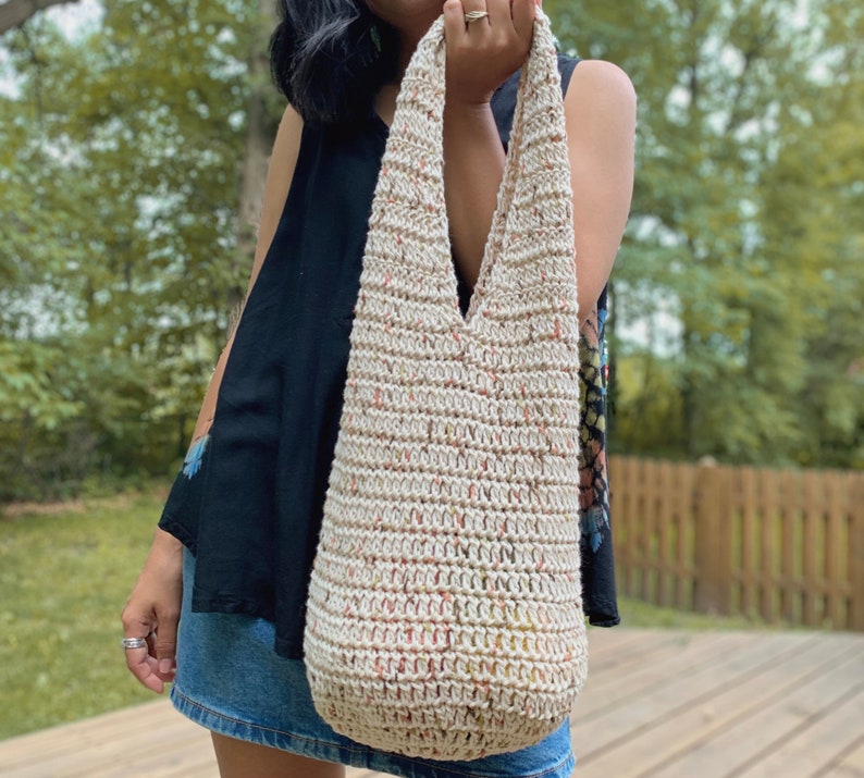 May include: A crocheted tote bag with a cream base and multi-colored flecks. The bag has a wide, v-shaped opening and a long strap.