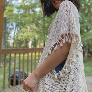 May include: A white crochet cardigan with a multicolored speckled pattern and fringe trim. The cardigan has a loose fit and is open in the front.