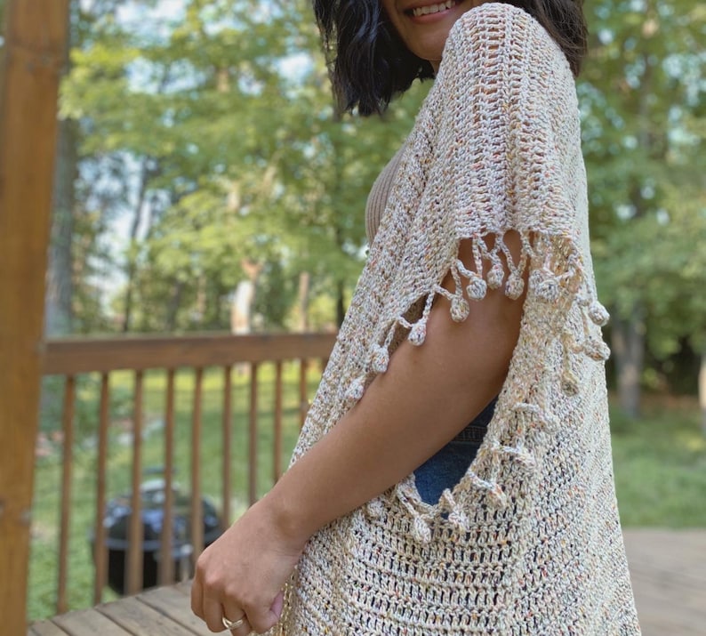 May include: A white crochet cardigan with a multicolored speckled pattern and fringe trim. The cardigan has a loose fit and is open in the front.