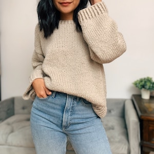 May include: A beige knitted sweater with a relaxed fit and long sleeves. The sweater is made of a soft, chunky yarn and has a slightly oversized look.