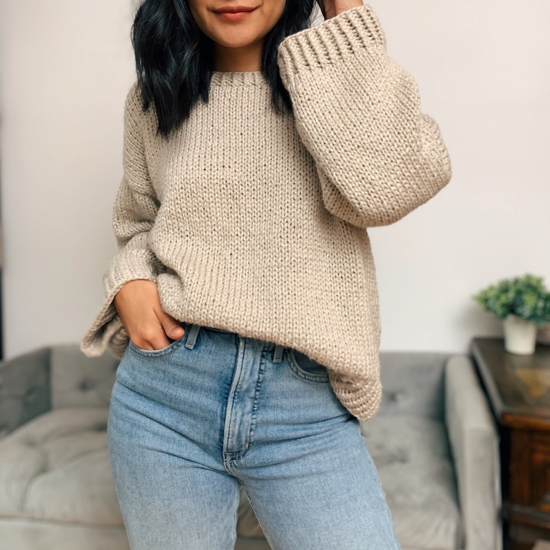 May include: A beige knitted sweater with a relaxed fit and long sleeves. The sweater is made of a soft, chunky yarn and has a slightly oversized look.