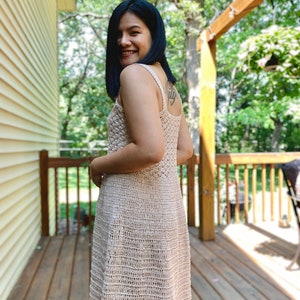 May include: A woman wearing a beige crochet dress with a sleeveless design. The dress has a fitted bodice and a flowing skirt. The woman is standing on a wooden deck with a green background.