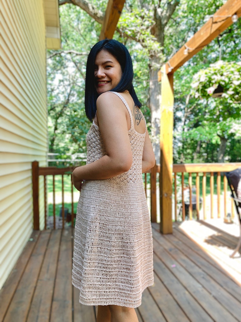 May include: A woman wearing a beige crochet dress with a sleeveless design. The dress has a fitted bodice and a flowing skirt. The woman is standing on a wooden deck with a green background.
