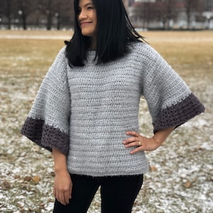 May include: A gray crocheted sweater with a darker gray trim on the sleeves. The sweater has a relaxed fit and a slightly cropped length.