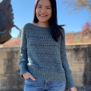 May include: A woman wearing a blue crocheted sweater with long sleeves and a relaxed fit. She is smiling and has her hands in her pockets.