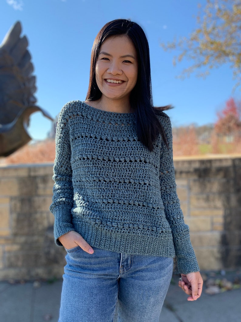 May include: A woman wearing a blue crocheted sweater with long sleeves and a relaxed fit. She is smiling and has her hands in her pockets.