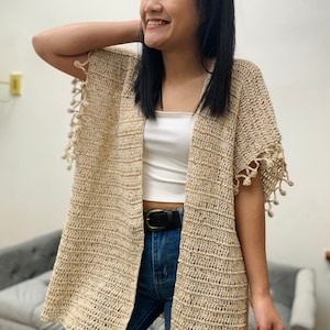 May include: A beige crochet cardigan with a speckled pattern and fringe detailing on the sleeves. The cardigan is open in the front and has a relaxed fit.