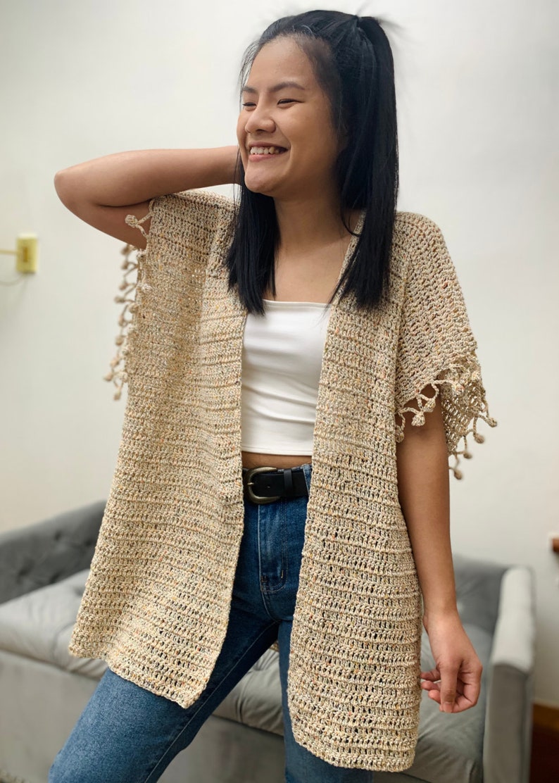 May include: A beige crochet cardigan with a speckled pattern and fringe detailing on the sleeves. The cardigan is open in the front and has a relaxed fit.