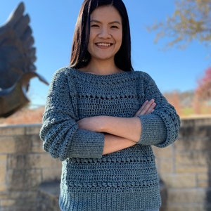 May include: A woman wearing a blue crocheted sweater with a loose fit and long sleeves. She is standing in front of a stone wall with a sculpture in the background.