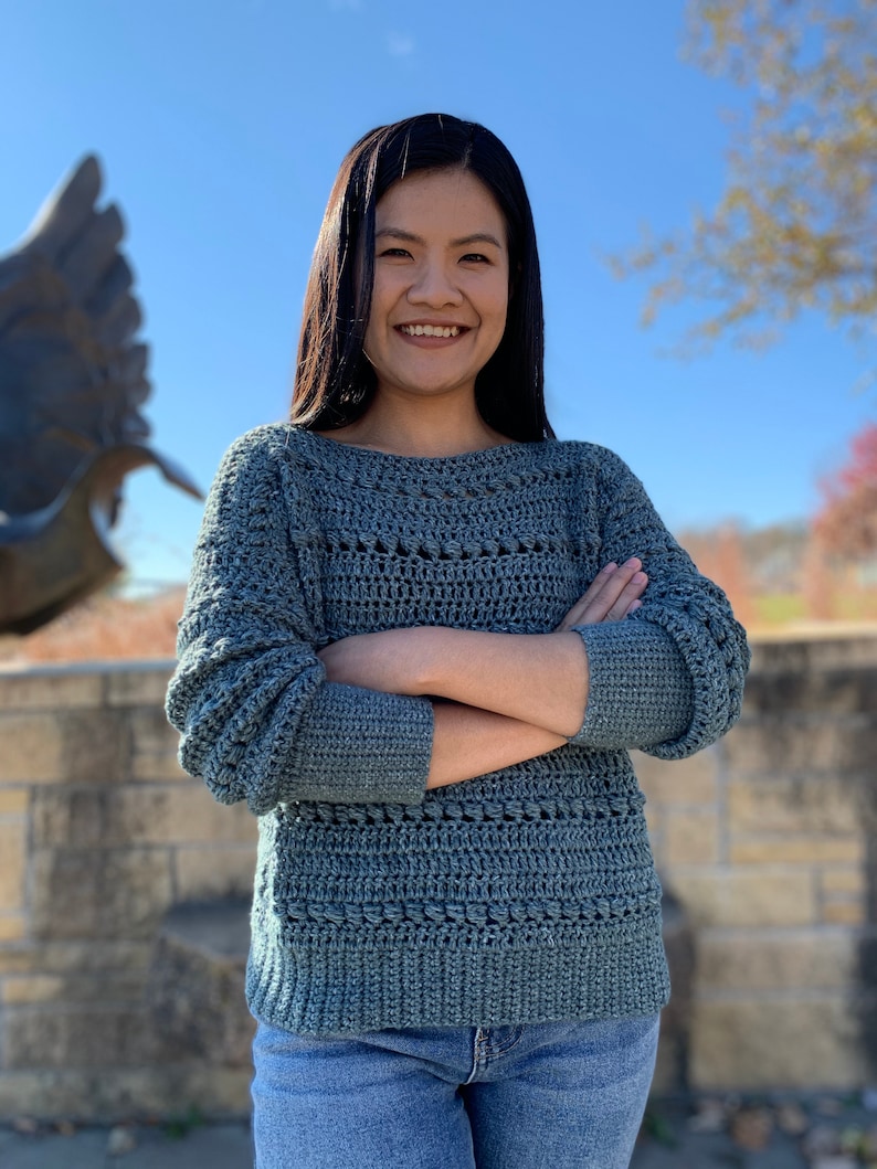 May include: A woman wearing a blue crocheted sweater with a loose fit and long sleeves. She is standing in front of a stone wall with a sculpture in the background.