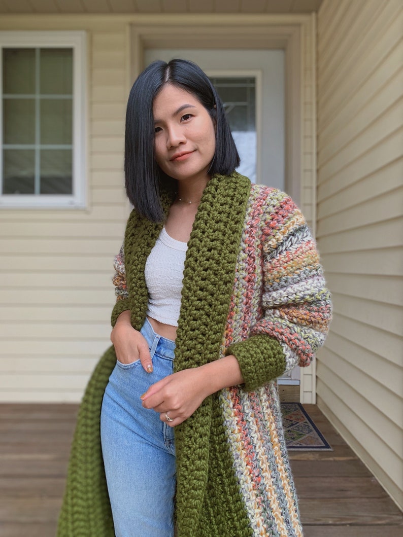 May include: A long, striped crochet cardigan with a green crocheted collar and sleeves. The cardigan is worn over a white tank top and blue jeans.