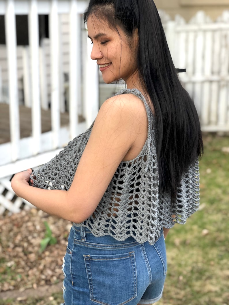May include: A grey crochet top with a scalloped hem. The top is sleeveless and has a loose fit. The model is wearing the top with denim shorts.