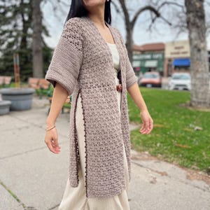 May include: A long, open-front, crochet cardigan in a neutral taupe colour. The cardigan features short sleeves and a detailed stitch pattern. The edges are trimmed with a darker brown border. The garment is worn over a cream-coloured dress.