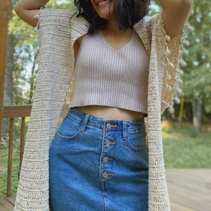May include: A woman wearing a cream crochet cardigan with a light pink tank top and a blue denim skirt. The cardigan has a fringe detail and the skirt has a button closure.