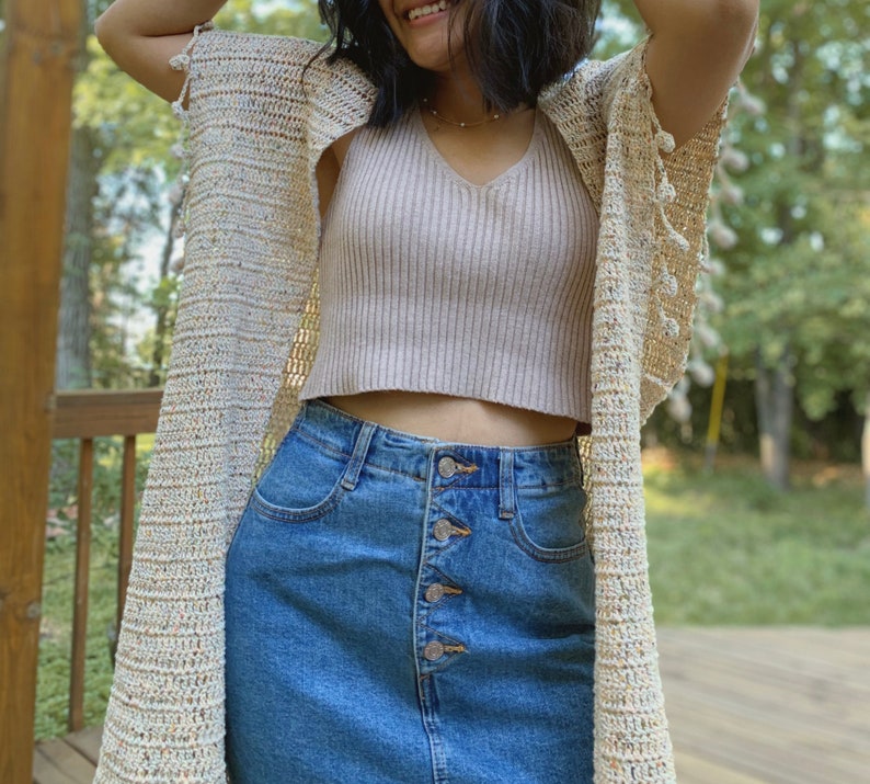 May include: A woman wearing a cream crochet cardigan with a light pink tank top and a blue denim skirt. The cardigan has a fringe detail and the skirt has a button closure.