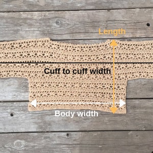 Puede incluir: Un top de ganchillo beige de tejido abierto, colocado plano sobre una superficie de madera. El top se mide con l&iacute;neas negras y amarillas que indican la longitud, el ancho de pu&ntilde;o a pu&ntilde;o y el ancho del cuerpo. El dise&ntilde;o de ganchillo presenta un patr&oacute;n delicado y aireado.