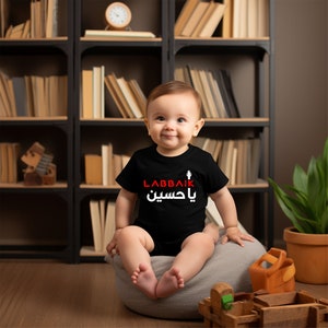 May include: A baby wearing a black onesie with the Arabic text "Labbaik Ya Hussein" in red and white. The baby is sitting on a gray beanbag chair in front of a bookshelf.