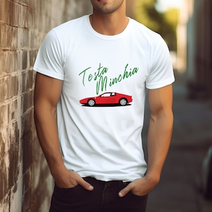 May include: White t-shirt featuring a red sports car graphic and the words "Testa Minchia" in green script. The car design is a side profile of a classic model. The shirt is made of a soft, comfortable material.