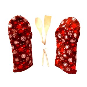 Könnte beinhalten: Ein Paar rote Ofenhandschuhe mit einem Schneeflockenmuster. Die Handschuhe sind aus Stoff und haben eine gesteppte Textur. Die Handschuhe liegen auf einer weißen Oberfläche neben zwei Holzlöffeln.