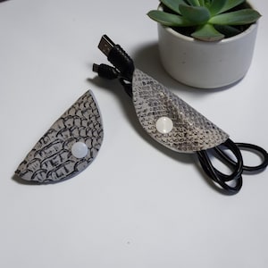 Könnte beinhalten: Zwei Kabelorganisatoren aus grau-weißem Kunstschlangenleder mit weißen Druckknöpfen. Ein Organizer hält ein schwarzes USB-Kabel. Der andere Organizer ist abgetrennt. Eine kleine Sukkulentenpflanze in einem weißen Topf befindet sich im Hintergrund.