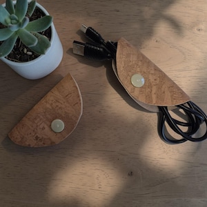 Könnte beinhalten: Zwei Kabelorganisatoren aus Kork mit hellem Knopfverschluss. Ein Organizer enthält schwarze Kabel. Eine kleine Sukkulentenpflanze in einem weißen Topf befindet sich im Hintergrund. Die Organizer befinden sich auf einer Holzoberfläche.