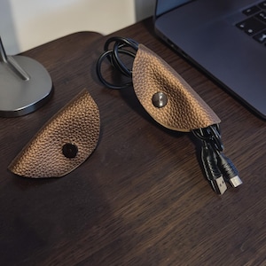Könnte beinhalten: Zwei braune Kabelorganisatoren aus Leder mit Druckknöpfen. Ein Organizer enthält schwarze Kabel und USB-Anschlüsse. Die Organizer befinden sich auf einer dunklen Holzoberfläche neben einem Laptop.
