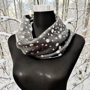 Könnte beinhalten: Ein grauer Loop-Schal mit weißen Punkten wird auf einer schwarzen Schaufensterpuppe präsentiert. Der Schal besteht aus einem weichen Material und ist zum Tragen um den Hals konzipiert. Der Hintergrund zeigt einen verschneiten Wald.