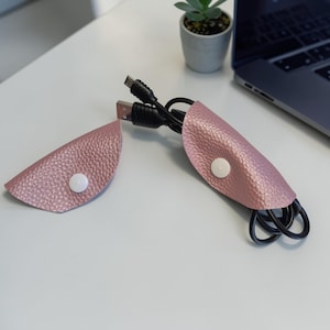 Könnte beinhalten: Zwei rosa Kabelorganisatoren aus Leder mit weißen Druckknöpfen. Ein Organizer enthält schwarze USB-Kabel. Die Organizer befinden sich auf einer weißen Oberfläche, mit einem Laptop und einer kleinen Topfpflanze im Hintergrund.