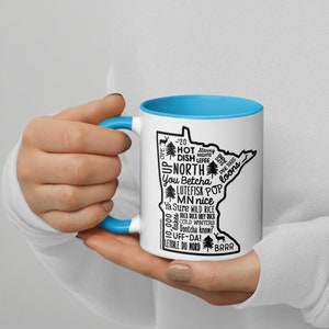 May include: A white ceramic mug with a light blue rim. The mug features a black outline of the state of Minnesota with text inside the outline. The text includes "You Betcha", "Lutefisk", "MN nice", "Wild Rice", "Cold Winters", "Dontcha know?", "UFF-DA!" and "Letoile du Nord".
