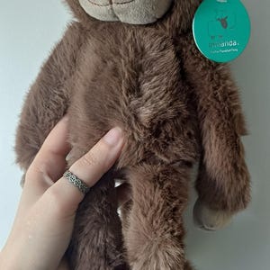 Peut inclure: Un singe en peluche marron avec un visage souriant et une fourrure douce. La peluche porte une étiquette indiquant "iwanda. softerThanASoftThing". Le singe est tenu par une personne.
