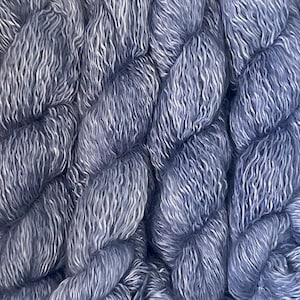 Puede incluir: Primer plano de un ovillo de hilo teñido a mano en tonos azules y blancos. El hilo es grueso y texturizado, con apariencia trenzada. El degradado de color crea un aspecto suave y etéreo, perfecto para proyectos de punto o ganchillo.