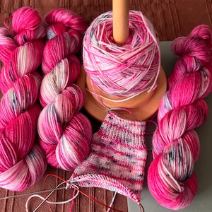 Könnte beinhalten: Drei Garnstränge in den Farben Pink, Weiß und dunkelblauen Sprenkeln, neben einer hölzernen Garnwinde mit einem Knäuel aus demselben Garn und einem teilweise gestrickten Stück auf Rundstricknadeln mit roten Seilen.