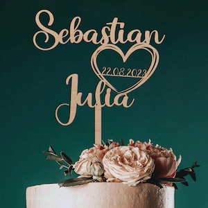 Op de afbeelding: Een bruidstaart met twee lagen, versierd met roze en crèmekleurige rozen en groen. Een houten taarttopper toont de namen "Sebastian" en "Julia", een hart en de datum "22.08.2023". De achtergrond is donkergroen.