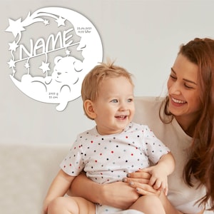 May include: A white wooden moon-shaped cutout with stars and a sleeping bear. The cutout includes the text "NAME", "22.06.2021", "11:15 Uhr", "2055 g", and "53 cm". A mother and child are smiling in the background.