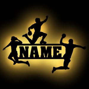 Könnte beinhalten: Schwarze Silhouette von drei Handballspielern, die mit einem Ball in der Luft springen. Die Spieler befinden sich über einem schwarzen Banner mit dem Wort "NAME" in weißen Buchstaben.