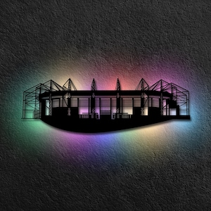 Puede incluir: Silueta negra de un estadio sobre un fondo texturizado. El estadio está iluminado con un arcoíris de colores, incluyendo verde, morado y rosa. El estadio presenta una estructura de techo detallada y áreas de asientos.